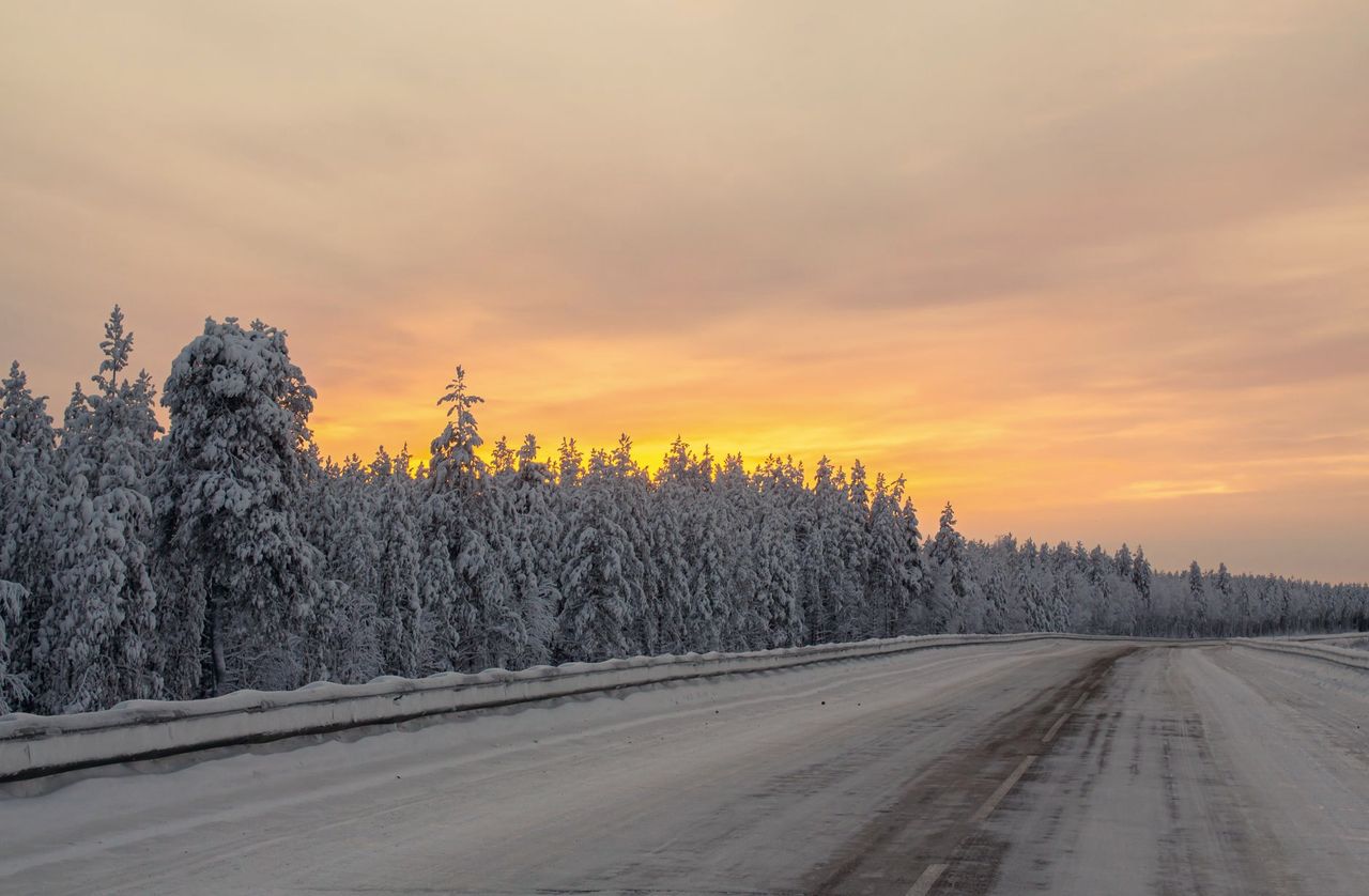 Route enneigée longeant une forêt givrée sous un ciel de coucher de soleil
